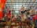A variety of colorful bags and fabrics are hanging from a ceiling grid in a shop. The ceiling has ropes and hooks holding the items. Below are wooden cabinets with more textiles and accessories displayed. The shop is vibrant with an assortment of patterns and colors.