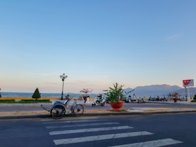 A scenic coastal road features a parked tricycle rickshaw on the right side, facing a sidewalk with people walking and riding scooters. The ocean is visible beyond the sidewalk, with gentle waves and a mountain range in the distance under a clear sky. Potted plants and street lamps line the walkway, adding a touch of greenery.