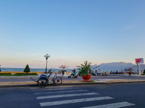 A scenic coastal road features a parked tricycle rickshaw on the right side, facing a sidewalk with people walking and riding scooters. The ocean is visible beyond the sidewalk, with gentle waves and a mountain range in the distance under a clear sky. Potted plants and street lamps line the walkway, adding a touch of greenery.