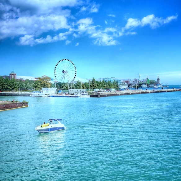 A serene waterfront scene with a small motorboat gliding through calm blue waters. In the background, a large Ferris wheel stands prominently against a bright blue sky dotted with a few clouds. The shoreline is lined with trees and buildings, including a dock and several moored boats. The overall atmosphere is peaceful and leisurely.