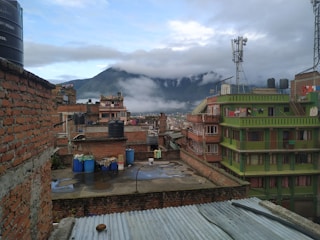 Urban rooftop view with multiple brick and concrete buildings. The scene includes a large number of water storage tanks and barrels on the rooftops. There is a green building with laundry hanging outside its windows. The background features mountains partially covered in clouds and mist, creating a serene atmosphere.