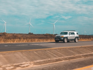 A silver SUV travels on a highway with a backdrop of several wind turbines in a rural setting. The road has visible lane markings, and the surroundings suggest open fields and a sky partly covered with clouds.