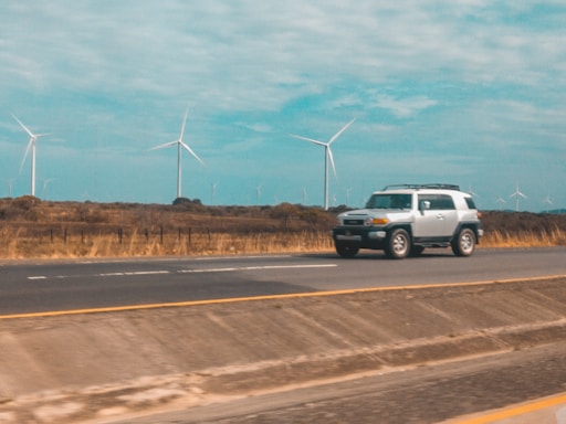 A silver SUV travels on a highway with a backdrop of several wind turbines in a rural setting. The road has visible lane markings, and the surroundings suggest open fields and a sky partly covered with clouds.