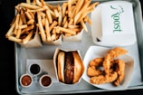 An overhead view of a tray filled with fast food items, including two boxes of golden fries, a cheeseburger in a wrapper, crispy chicken tenders in a box, and small containers of dipping sauces. The words 'Roost' are visible on one of the takeout boxes.