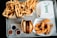 An overhead view of a tray filled with fast food items, including two boxes of golden fries, a cheeseburger in a wrapper, crispy chicken tenders in a box, and small containers of dipping sauces. The words 'Roost' are visible on one of the takeout boxes.