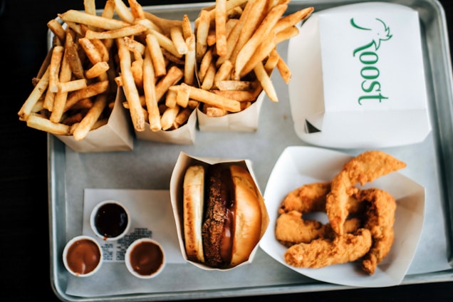 An overhead view of a tray filled with fast food items, including two boxes of golden fries, a cheeseburger in a wrapper, crispy chicken tenders in a box, and small containers of dipping sauces. The words 'Roost' are visible on one of the takeout boxes.