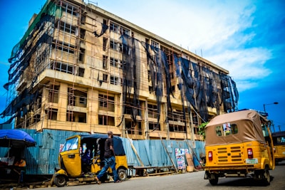 A multi-story building under construction is covered with scaffolding and black protective netting. In the foreground, a street scene includes two yellow auto rickshaws, one moving away and the other parked with people inside. A person is walking nearby and the sky above is clear with a few clouds.