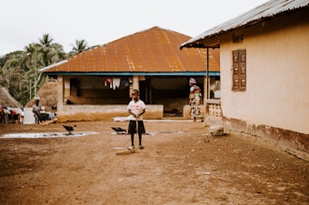 A young child stands in a dusty village area holding a stick, surrounded by traditional houses with rusty metal roofs. In the background, a woman dressed in colorful clothing and another person can be seen near the entrance of one of the homes. Chickens roam freely on the ground, and there are palm trees in the distance, contributing to a rural setting.