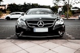 A sleek black Mercedes-Benz car is parked on a tiled pavement, with a focus on the front end showing the iconic emblem on the grille. The background features other parked vehicles, greenery, and some residential buildings.