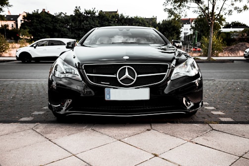 A sleek black Mercedes-Benz car is parked on a tiled pavement, with a focus on the front end showing the iconic emblem on the grille. The background features other parked vehicles, greenery, and some residential buildings.