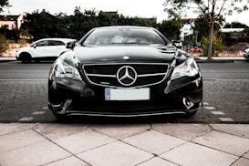 A sleek black Mercedes-Benz car is parked on a tiled pavement, with a focus on the front end showing the iconic emblem on the grille. The background features other parked vehicles, greenery, and some residential buildings.