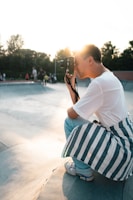 A person holding a camera is crouched down, capturing a moment at a skate park during sunset. The person is dressed casually, with a white shirt and striped jacket tied around the waist. The background features skaters enjoying the park, and trees are visible under a bright sky.