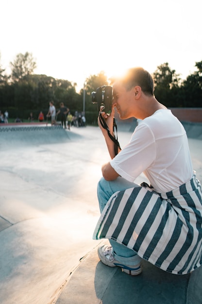 A person holding a camera is crouched down, capturing a moment at a skate park during sunset. The person is dressed casually, with a white shirt and striped jacket tied around the waist. The background features skaters enjoying the park, and trees are visible under a bright sky.