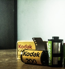 Two film rolls, Kodak and Fujicolor, are positioned on a wooden surface with a Kodak film box in the background. The Kodak film roll is yellow with black text, while the Fujicolor roll is green with white text. A short strip of film protrudes from the Kodak roll.