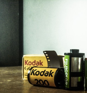 Two film rolls, Kodak and Fujicolor, are positioned on a wooden surface with a Kodak film box in the background. The Kodak film roll is yellow with black text, while the Fujicolor roll is green with white text. A short strip of film protrudes from the Kodak roll.