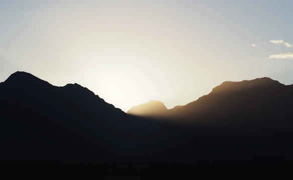 A serene landscape of silhouetted mountains against a bright, rising sun. The sky features a gradient from deep blue at the top to a soft yellow near the horizon, where the sunlight peeks through the peaks.