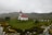 A small church with a red roof and white walls stands in a green field surrounded by a cemetery with numerous headstones. The background features rolling hills under a cloudy sky, contributing to a serene and somber atmosphere.