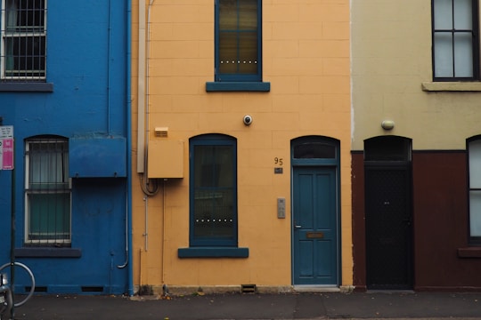 An image of an urban street view featuring the exteriors of three distinct houses. The house in the center is painted yellow with blue trim around the door and windows, marked with the number 95. The houses on either side are painted blue and a muted brown, respectively. Various details such as windows, doors, a street sign, and part of a bicycle are visible.