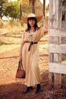 A woman stands outdoors in a rustic setting. She is wearing a long beige dress with embroidered details, a wide-brimmed straw hat, and black boots. She carries a brown leather handbag and is leaning against a weathered wooden structure. The background consists of trees and dry, earthy ground.