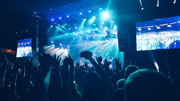 A vibrant concert scene with a large crowd of people raising their hands in the air. Bright blue stage lights illuminate the venue, with a band performing on stage. Large screens display live footage of the audience, creating an immersive atmosphere.