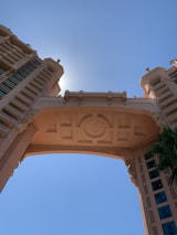 A detailed architectural structure featuring a large, ornate archway flanked by two towering buildings. The design includes intricate geometric patterns and appears to be bathed in sunlight, with the sun positioned directly behind the central arch. The sky is clear and blue, enhancing the structure’s grandeur.