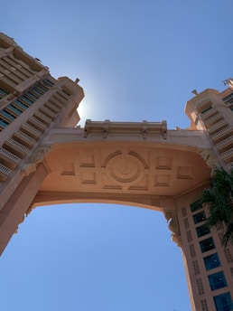 A detailed architectural structure featuring a large, ornate archway flanked by two towering buildings. The design includes intricate geometric patterns and appears to be bathed in sunlight, with the sun positioned directly behind the central arch. The sky is clear and blue, enhancing the structure’s grandeur.