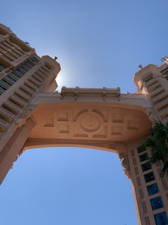 A detailed architectural structure featuring a large, ornate archway flanked by two towering buildings. The design includes intricate geometric patterns and appears to be bathed in sunlight, with the sun positioned directly behind the central arch. The sky is clear and blue, enhancing the structure’s grandeur.