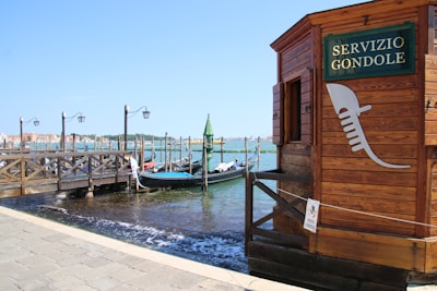 A wooden gondola service booth is situated by a canal with several gondolas moored alongside a wooden dock. The booth bears a sign that reads 'SERVIZIO GONDOLE'. Bright blue sky and water set a serene backdrop, with traditional lampposts lining the dock.
