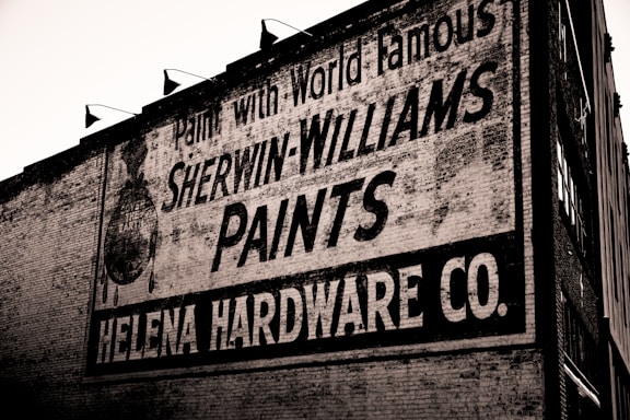 A brick wall displays a large vintage advertisement for Sherwin-Williams Paints and Helena Hardware Co. The painted sign is weathered, showing signs of age and wear, giving it an antique appearance. The text is bold and prominently positioned, with an iconic paint can logo visible on the left.