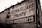 A brick wall displays a large vintage advertisement for Sherwin-Williams Paints and Helena Hardware Co. The painted sign is weathered, showing signs of age and wear, giving it an antique appearance. The text is bold and prominently positioned, with an iconic paint can logo visible on the left.