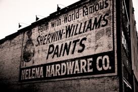 A brick wall displays a large vintage advertisement for Sherwin-Williams Paints and Helena Hardware Co. The painted sign is weathered, showing signs of age and wear, giving it an antique appearance. The text is bold and prominently positioned, with an iconic paint can logo visible on the left.