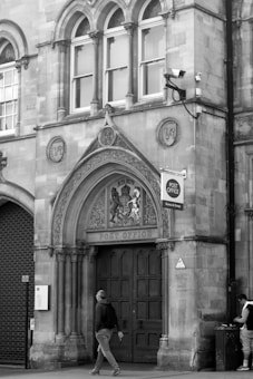 A historic building facade features ornate architectural details, including arched windows and decorative stonework. The entrance is marked by a large wooden door with the words 'Post Office' above it. A person walks past on the sidewalk, and another stands at the corner near a trash bin. A CCTV camera is mounted on the wall above the door.