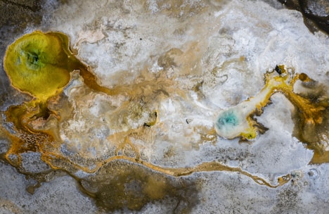 Aerial view of a geothermal landscape with vibrant mineral deposits. Dominant features include a bright yellow and green pool on the left with branching patterns extending across the surface. Pale blue and white mineral formations create a stark contrast with the muted brown and gray earth tones.