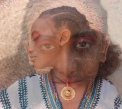 A surrealistic image featuring the overlapping faces of two individuals with braided hairstyles. The overlapping effect creates a dreamlike and abstract appearance. One individual is wearing a patterned garment and a sun-shaped pendant.