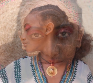 A surrealistic image featuring the overlapping faces of two individuals with braided hairstyles. The overlapping effect creates a dreamlike and abstract appearance. One individual is wearing a patterned garment and a sun-shaped pendant.