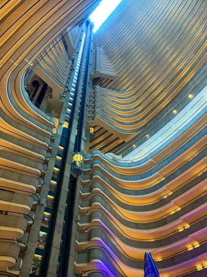 An interior view of a modern architectural space featuring multiple levels of balconies with curved designs, illuminated with warm lighting. A glass elevator is visible traveling between the floors, adding a sense of verticality and depth to the structure.