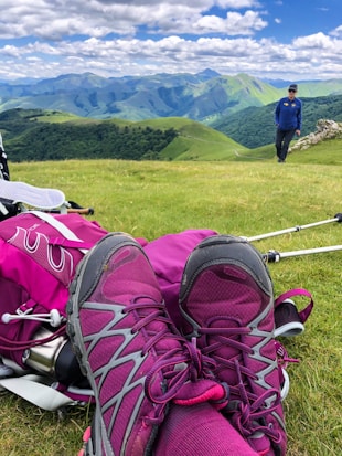 A pair of purple hiking boots are in the foreground against a backdrop of lush green hills and mountains. A person wearing a blue jacket and hat is walking in the middle distance. Hiking gear, including a pink backpack and trekking poles, is scattered around on the grass. The sky is bright with plentiful puffy clouds.