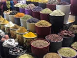 A vibrant display of various spices and herbs arranged in multiple large bowls. The containers are filled with colorful ingredients, such as dried peppers, turmeric, and other spices, creating a visually appealing and aromatic market scene.