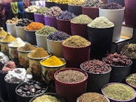 A vibrant display of various spices and herbs arranged in multiple large bowls. The containers are filled with colorful ingredients, such as dried peppers, turmeric, and other spices, creating a visually appealing and aromatic market scene.