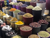 A vibrant display of various spices and herbs arranged in multiple large bowls. The containers are filled with colorful ingredients, such as dried peppers, turmeric, and other spices, creating a visually appealing and aromatic market scene.