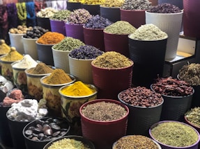 A vibrant display of various spices and herbs arranged in multiple large bowls. The containers are filled with colorful ingredients, such as dried peppers, turmeric, and other spices, creating a visually appealing and aromatic market scene.