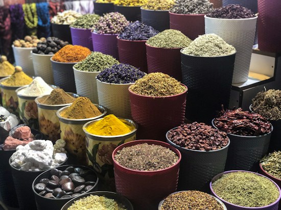 A vibrant display of various spices and herbs arranged in multiple large bowls. The containers are filled with colorful ingredients, such as dried peppers, turmeric, and other spices, creating a visually appealing and aromatic market scene.