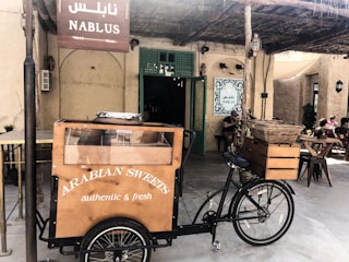 A bicycle cart selling Arabian sweets is positioned in front of an establishment adorned with a Nablus sign. The cart features a wooden display case with a glass front, and the words 'Arabian Sweets authentic & fresh' are printed on it. The setting includes a rustic outdoor area with straw roofing and a few diners seated at wooden tables, enjoying the ambiance.