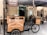 A bicycle cart selling Arabian sweets is positioned in front of an establishment adorned with a Nablus sign. The cart features a wooden display case with a glass front, and the words 'Arabian Sweets authentic & fresh' are printed on it. The setting includes a rustic outdoor area with straw roofing and a few diners seated at wooden tables, enjoying the ambiance.