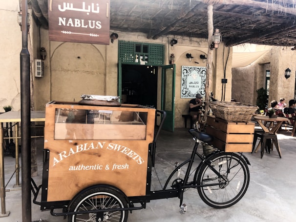 A bicycle cart selling Arabian sweets is positioned in front of an establishment adorned with a Nablus sign. The cart features a wooden display case with a glass front, and the words 'Arabian Sweets authentic & fresh' are printed on it. The setting includes a rustic outdoor area with straw roofing and a few diners seated at wooden tables, enjoying the ambiance.