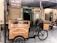 A bicycle cart selling Arabian sweets is positioned in front of an establishment adorned with a Nablus sign. The cart features a wooden display case with a glass front, and the words 'Arabian Sweets authentic & fresh' are printed on it. The setting includes a rustic outdoor area with straw roofing and a few diners seated at wooden tables, enjoying the ambiance.