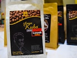 A package of Aman Kuba Luwak coffee is prominently displayed in the foreground, featuring a graphic of coffee beans and gold text on a black background. Other similar coffee packages are arranged in the background, slightly blurred.
