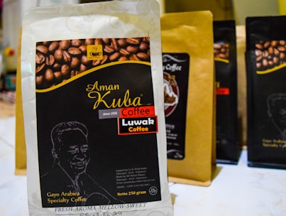 A package of Aman Kuba Luwak coffee is prominently displayed in the foreground, featuring a graphic of coffee beans and gold text on a black background. Other similar coffee packages are arranged in the background, slightly blurred.