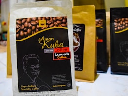 A package of Aman Kuba Luwak coffee is prominently displayed in the foreground, featuring a graphic of coffee beans and gold text on a black background. Other similar coffee packages are arranged in the background, slightly blurred.