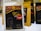 A package of Aman Kuba Luwak coffee is prominently displayed in the foreground, featuring a graphic of coffee beans and gold text on a black background. Other similar coffee packages are arranged in the background, slightly blurred.